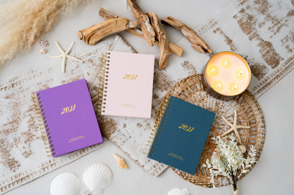 Val Marie Paper 2027 Rhythms prayer journals in Rosewater, Amethyst, and Dusk, styled with a candle, coffee, eucalyptus, and a cozy cream knit blanket