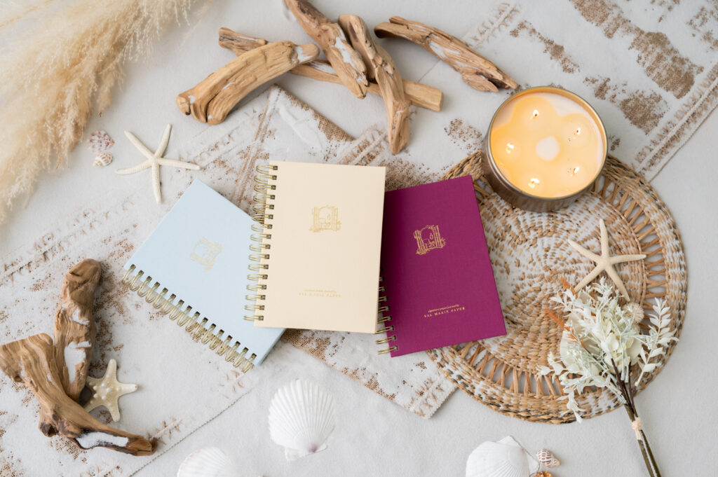 Val Marie Paper 2027 Signature prayer journals in Sky, Cerise, and Butter Yellow on a coastal flat lay with driftwood, seashells, starfish, and a candle